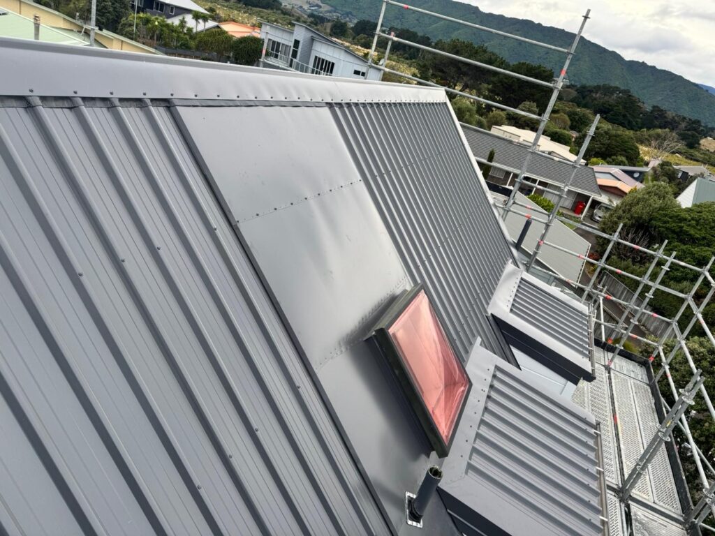 Dark long-run metal roof installation around a protected skylight with scaffolding visible