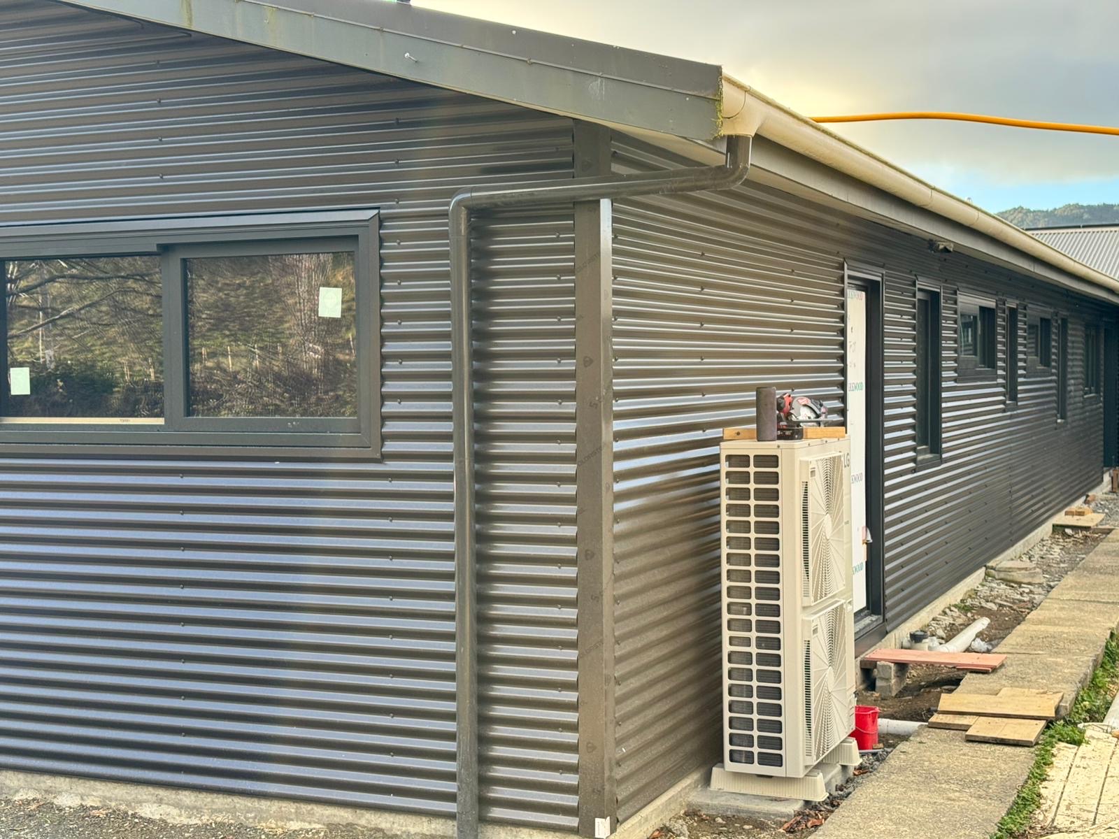 Dark horizontal metal wall cladding wrapping around a building corner beside a heat-pump unit